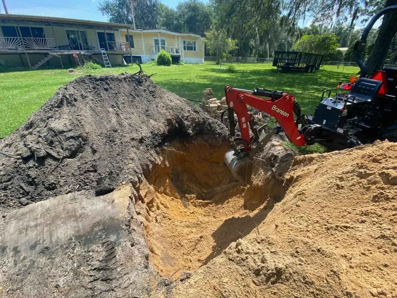 Red excavator digging a large hole beside dirt piles in a sunny yard near a house