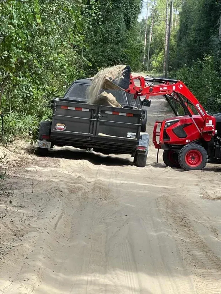 Red front loader dumping rocks into a black dump truck on a dirt road surrounded by trees