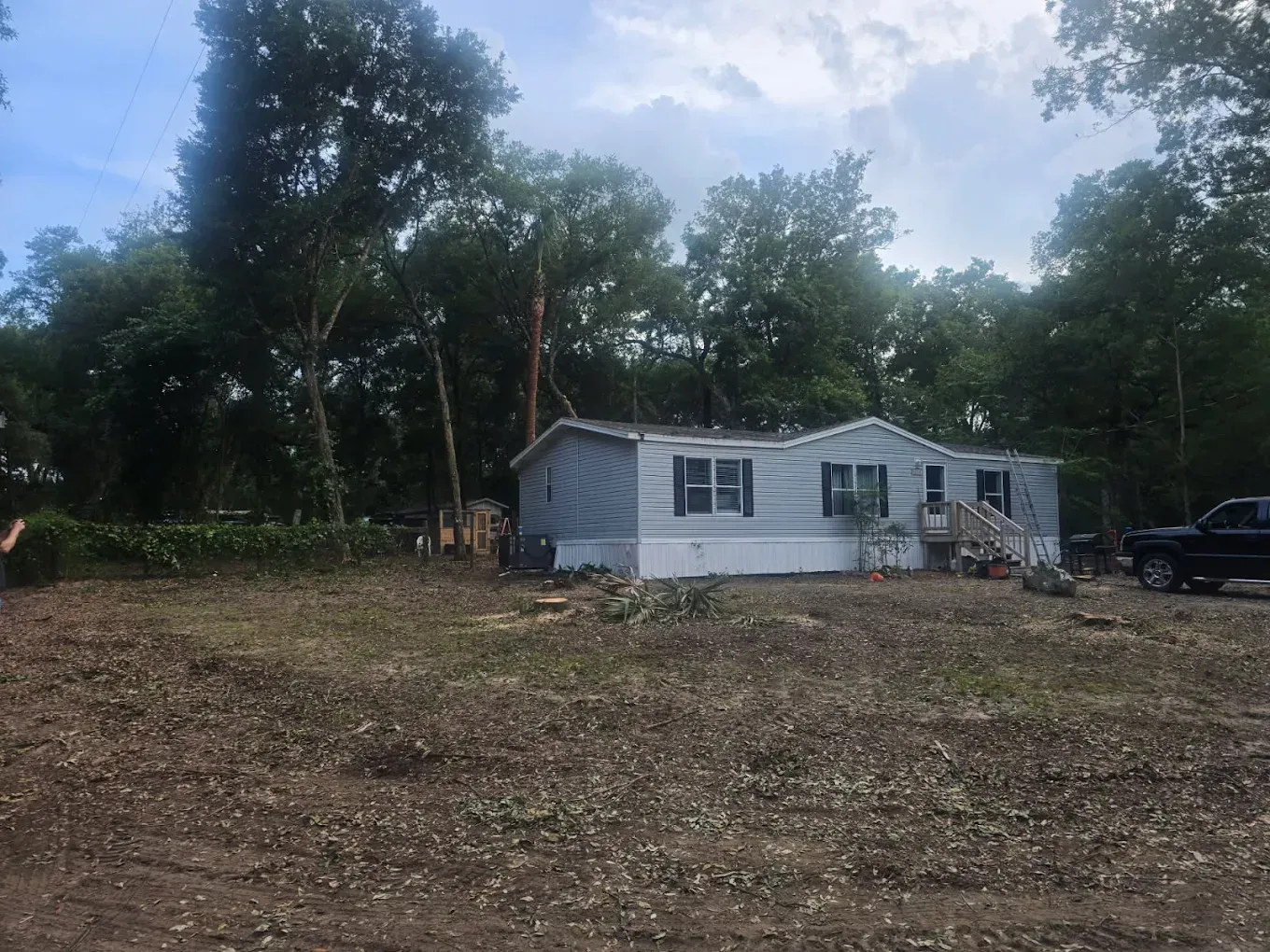 Small white house in a wooded clearing with a parked car and dirt foreground under cloudy sky