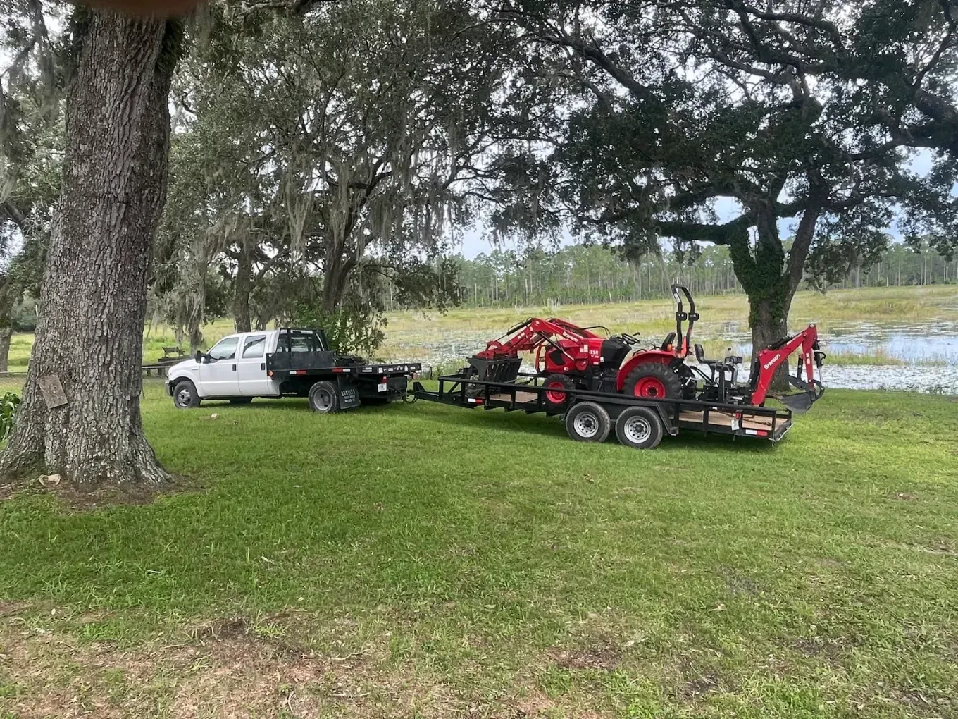 White pickup towing a red tractor on a trailer across a grassy field by a pond