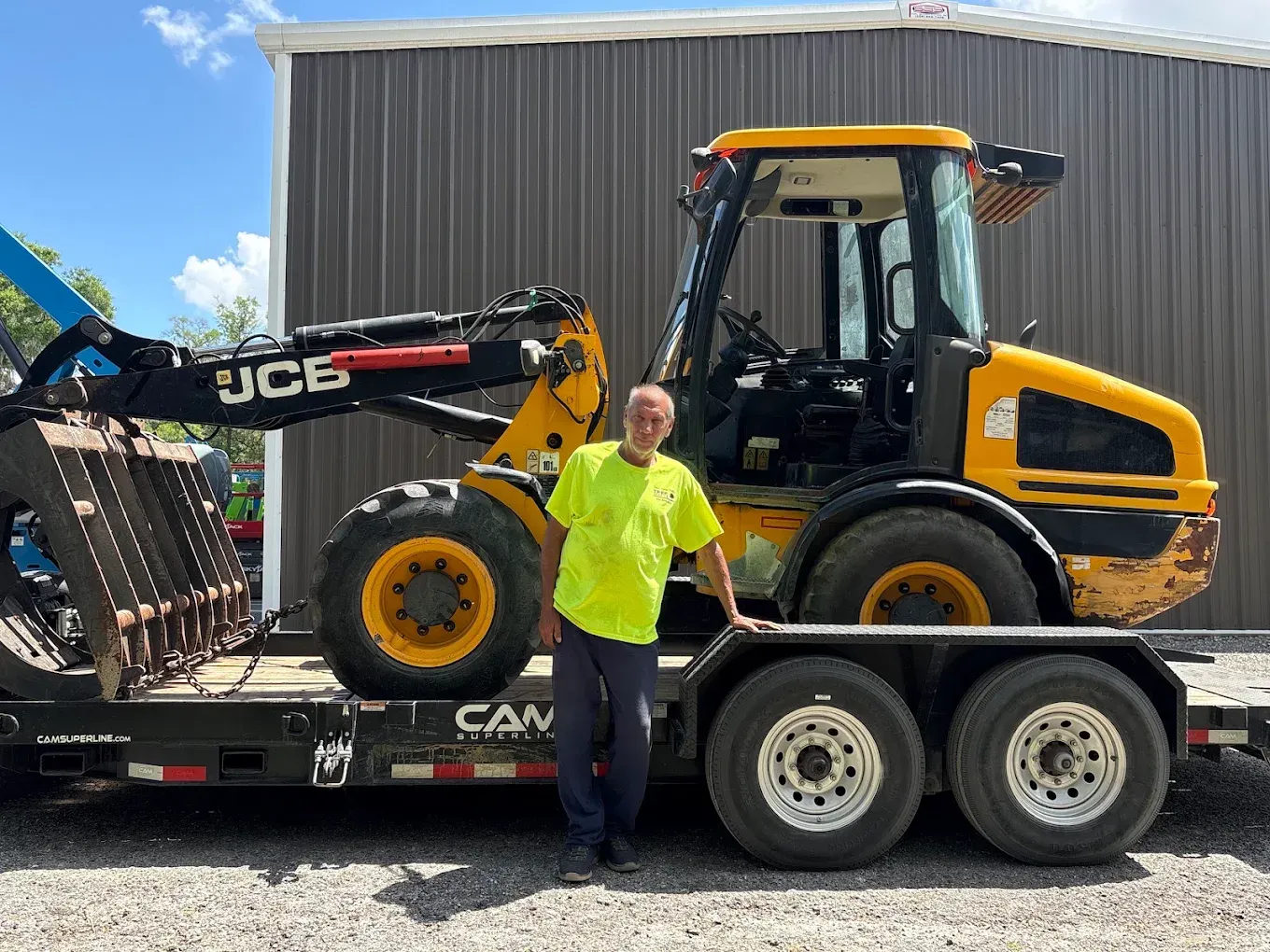 Yellow JCB loader on a trailer beside a person in a neon shirt, parked outdoors by a metal building
