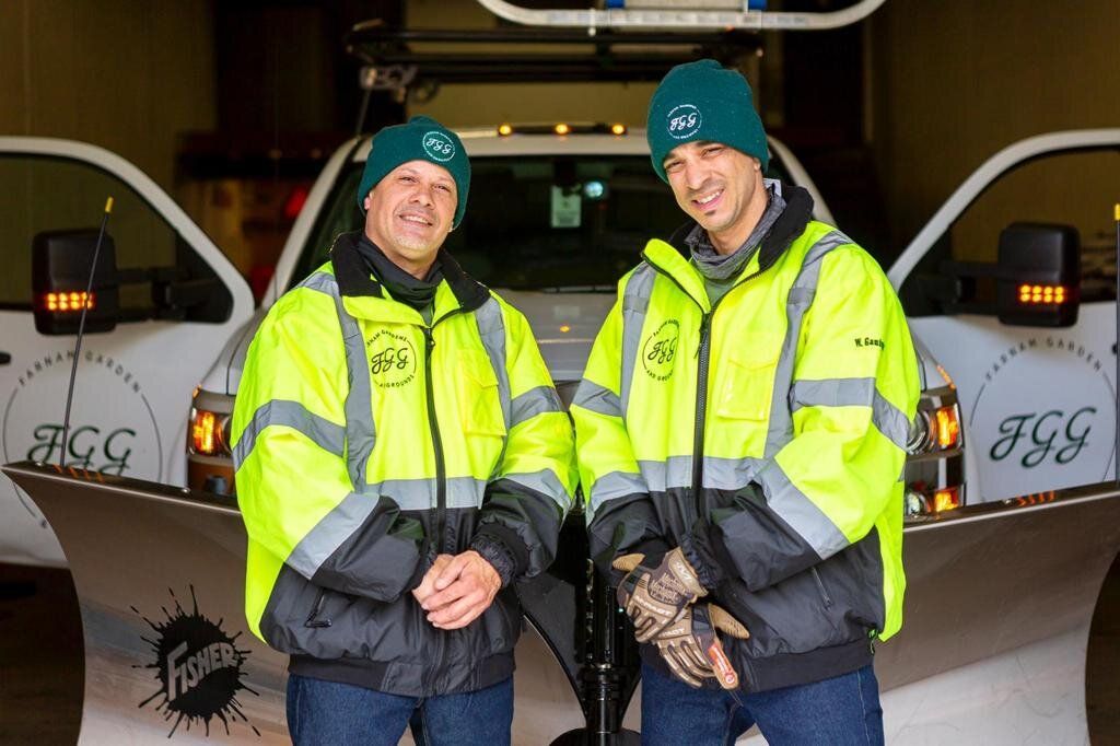 Two men are standing in front of a snow plow truck.