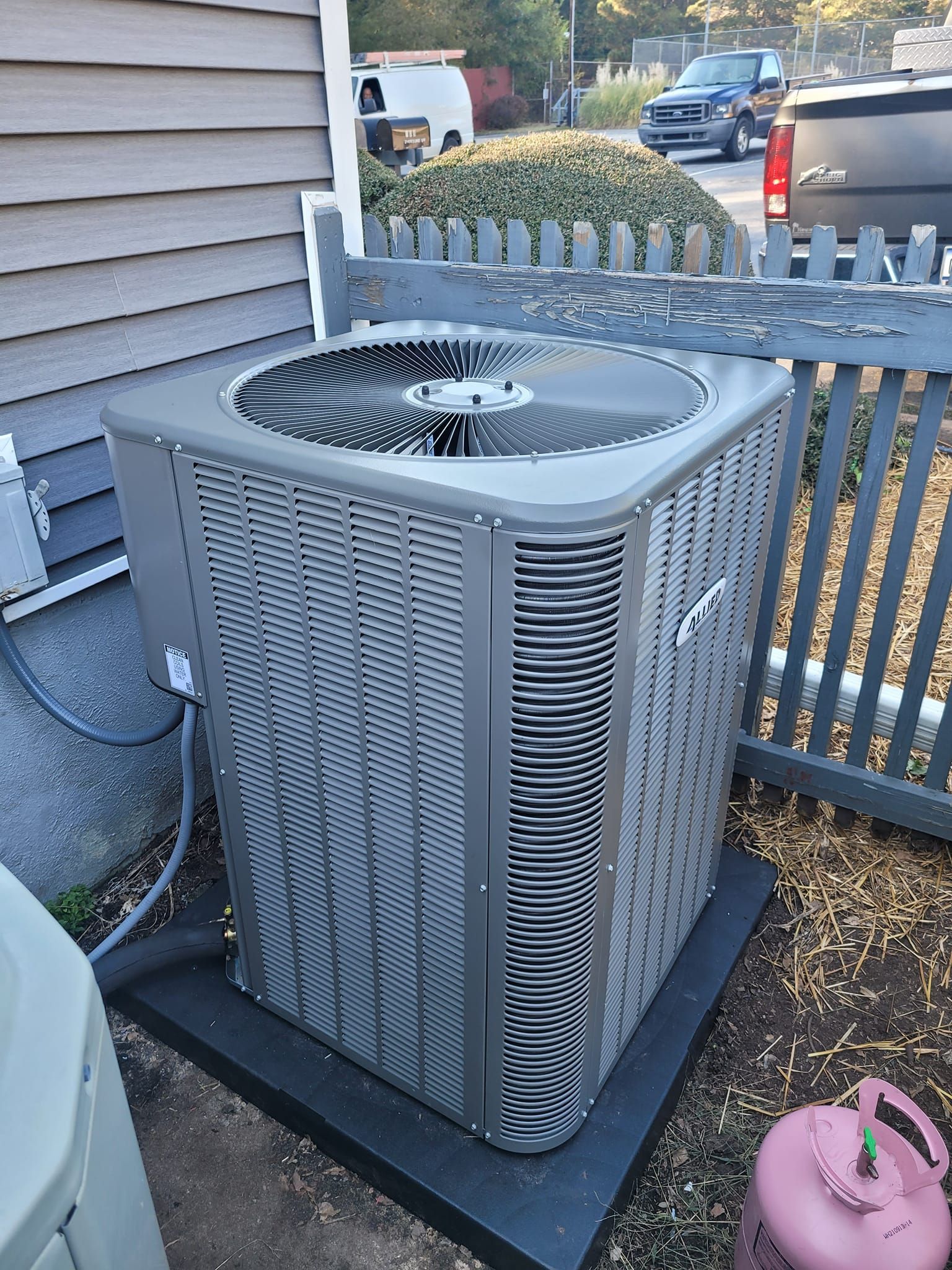 A large air conditioner is sitting outside of a house next to a fence.