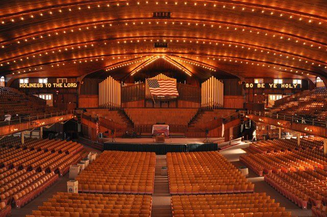 Interior of large auditorium with rows of seats, stage, and organ. Warm lighting, American flag.