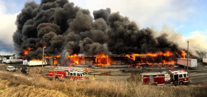 Fire engulfs a large building, with thick black smoke billowing. Fire trucks on the scene.