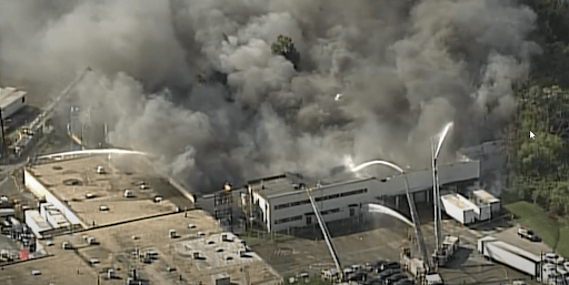 A large industrial building engulfed in flames and billowing thick smoke. Firefighters spray water from above.