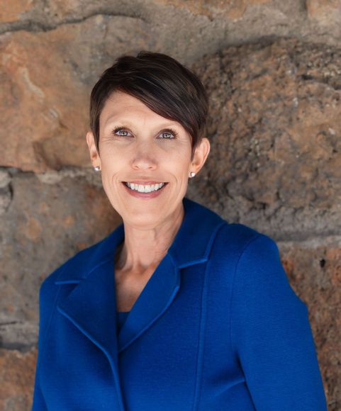 Woman with short dark hair wearing a blue jacket smiles in front of a stone wall.