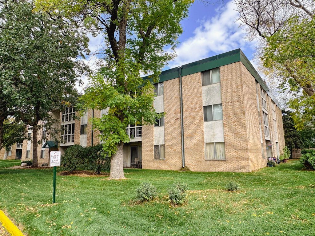 A large brick apartment building with a green roof is surrounded by trees and grass.