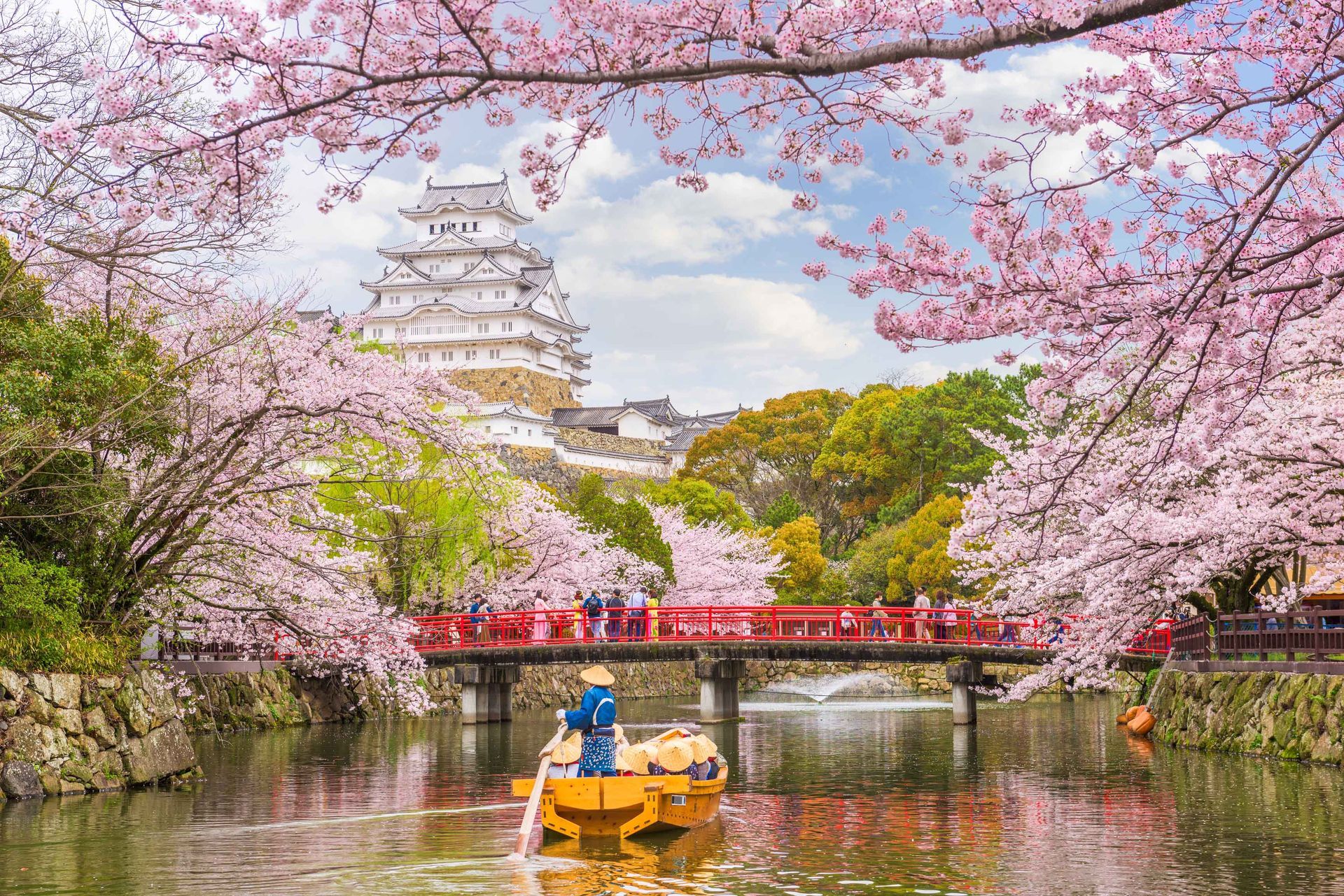 A tiny yellow boat sailing through a river in front of Himeji Castle in Japan, with pink trees enhancing the scenery.