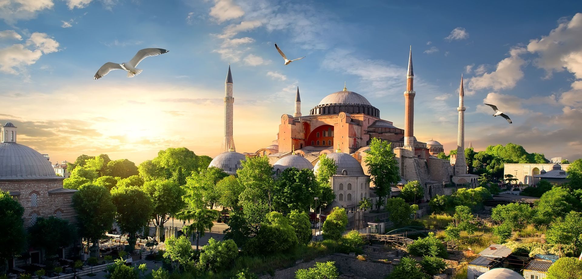 An aerial view of a mosque surrounded by trees and birds at sunset.