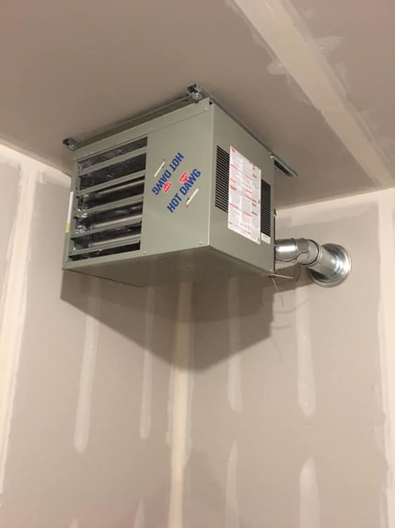 Ceiling-mounted, gray garage heater with exhaust pipe and warning labels in a room with drywall walls.