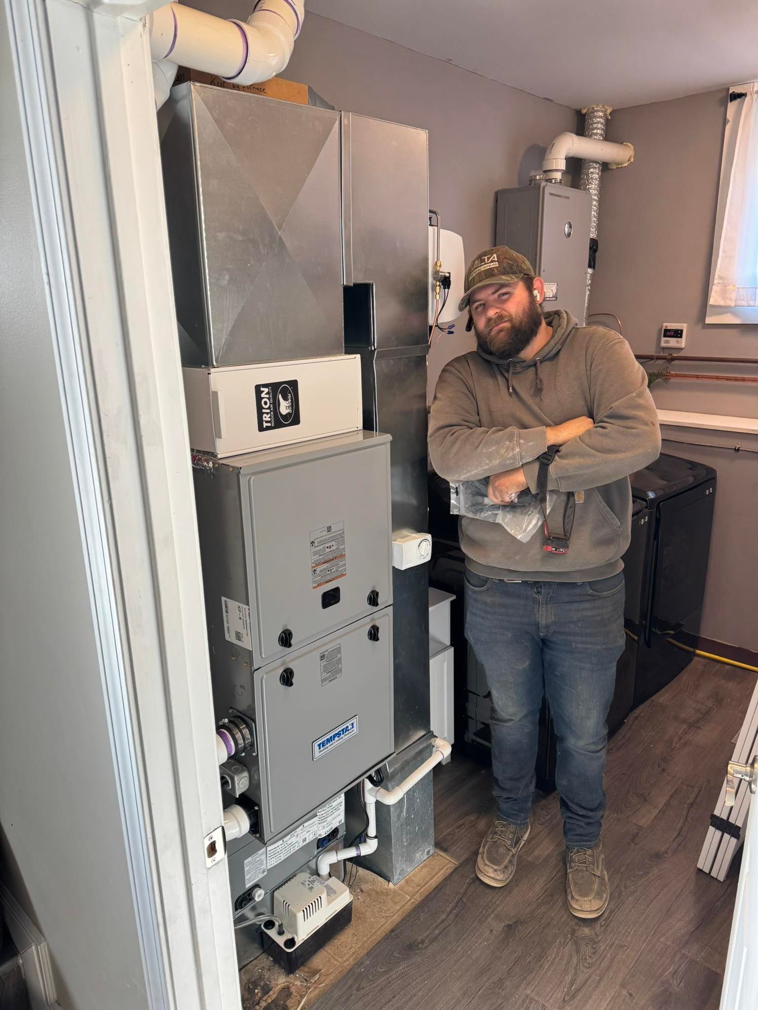 Man standing next to new furnace, arms crossed, in a utility room. Gray equipment and walls, brown flooring.