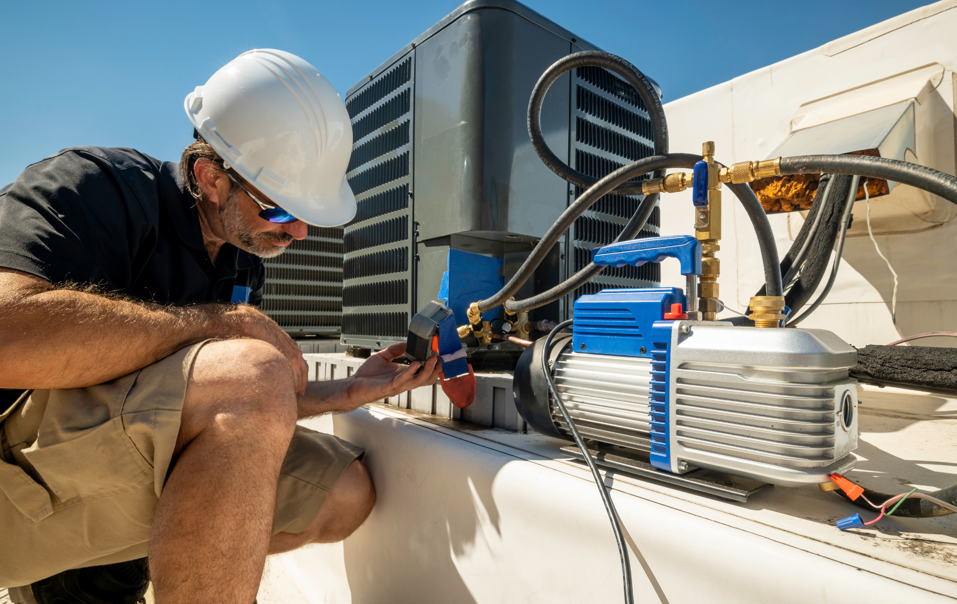 HVAC technician in a hard hat, working on rooftop equipment with vacuum pump. Sunny day.