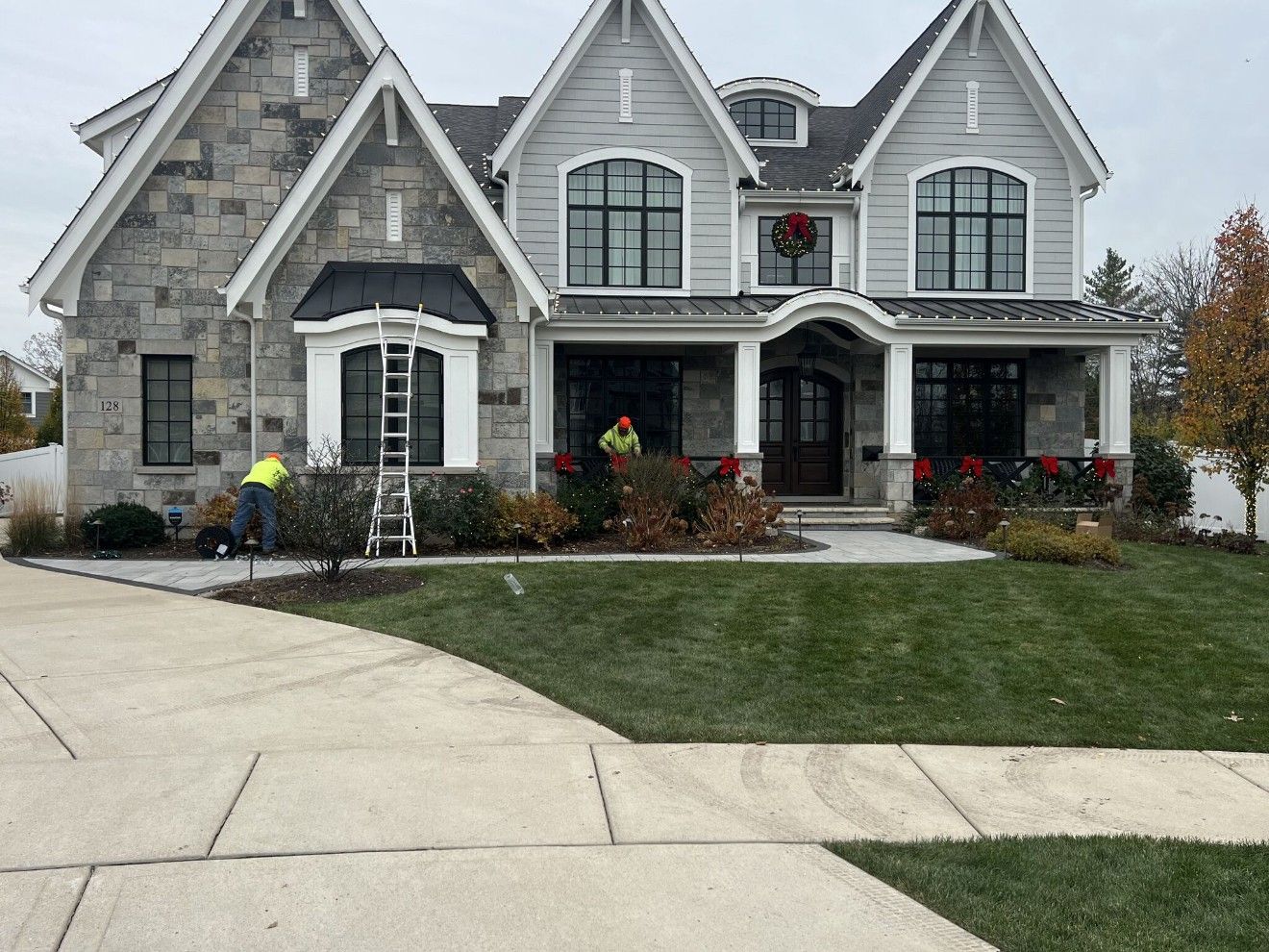 Gray house with stone facade, lawn, driveway. Workers on ladder installing holiday decorations.