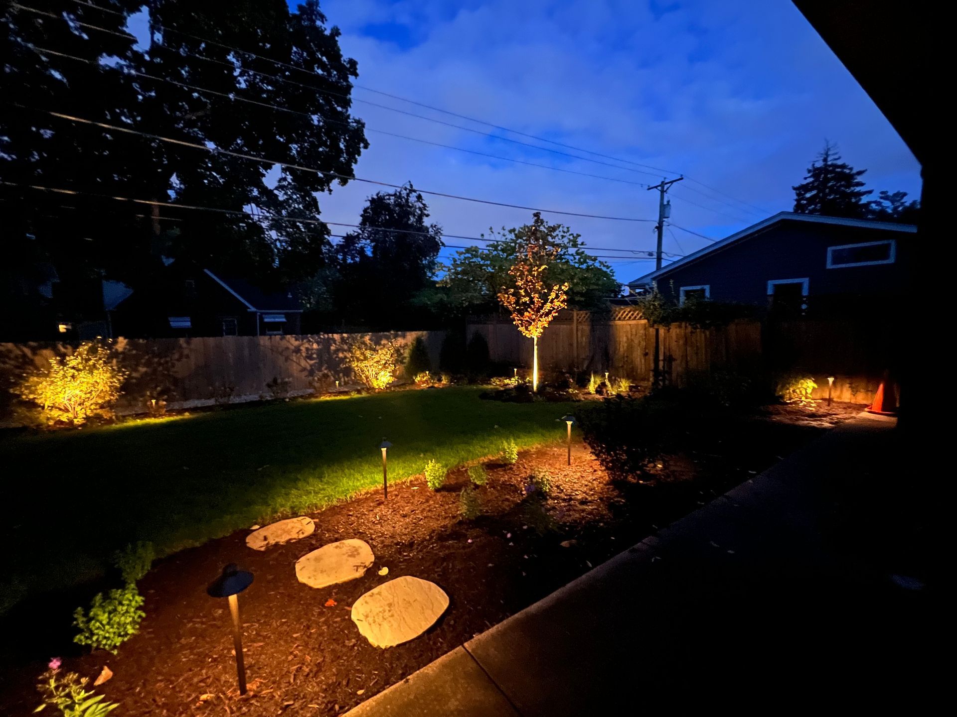 Nighttime backyard lit with landscape lights; grass, trees, and stepping stones.