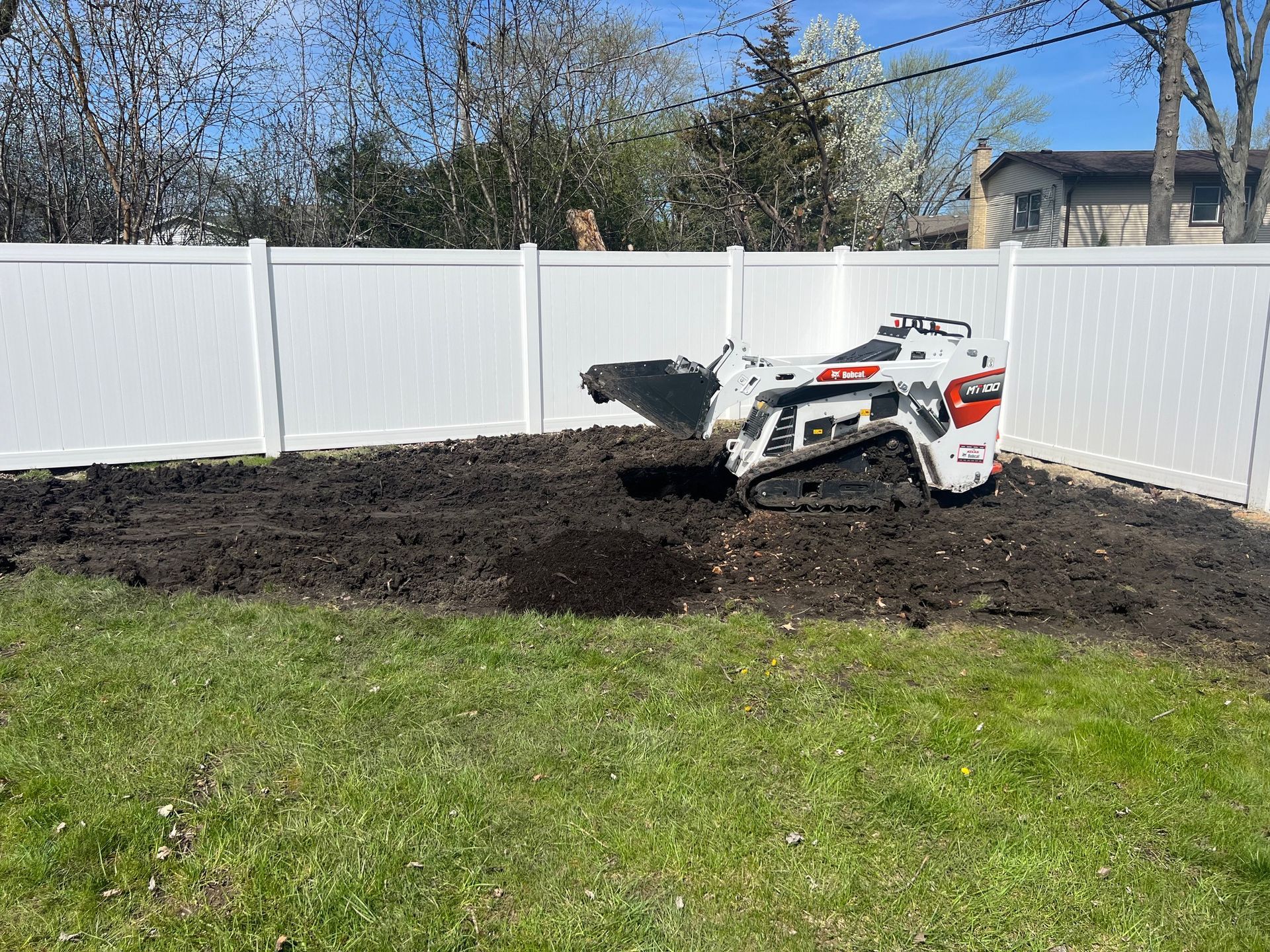 A stump grinder is sitting in a yard next to a white fence.