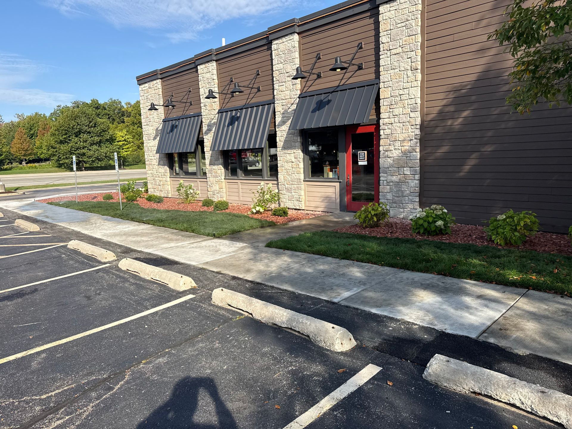 Exterior of a building with dark brick and stone accents, awnings, and a sidewalk along a parking lot.