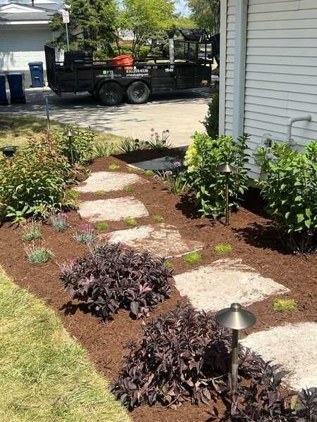 Stone pathway through a garden bed with dark purple plants and mulch. A trailer is in the background.