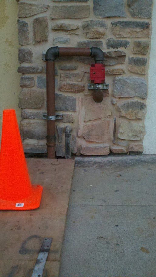 An orange cone is sitting next to a pipe in front of a stone wall.