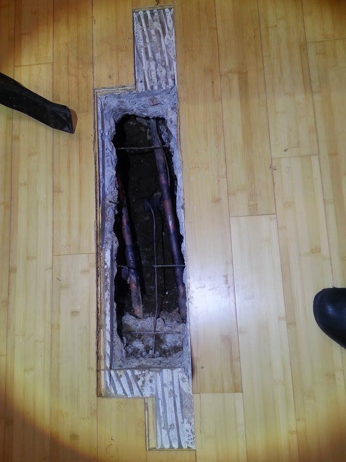 A hole in a wooden floor with a vacuum cleaner in the background.