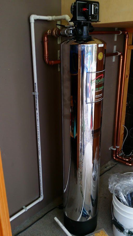 A stainless steel water filter is sitting in a room next to a bucket.