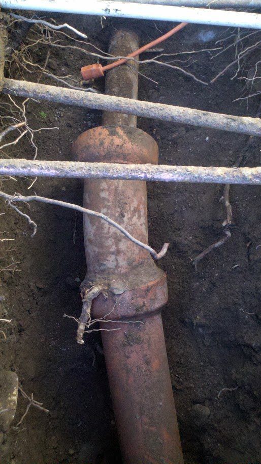 A rusty pipe is sitting in the dirt under a pipe.