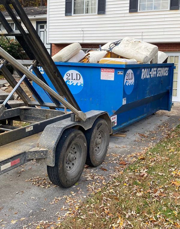 Blue dumpster on a trailer, filled with debris, parked on a driveway in front of a house.