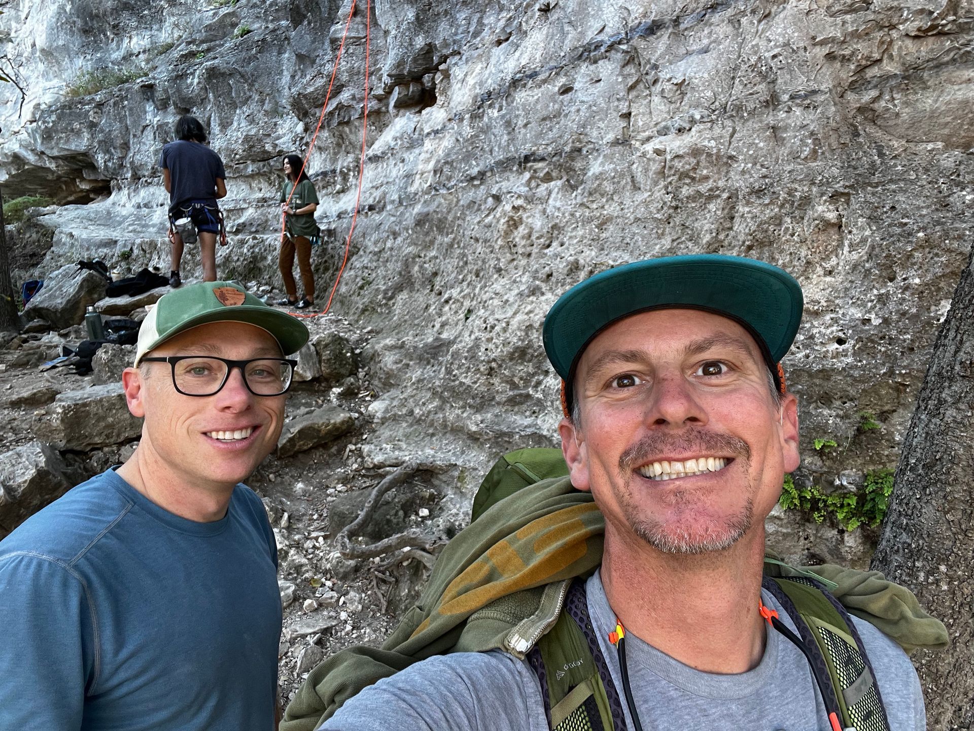 Two men are standing next to each other in front of a rock wall.