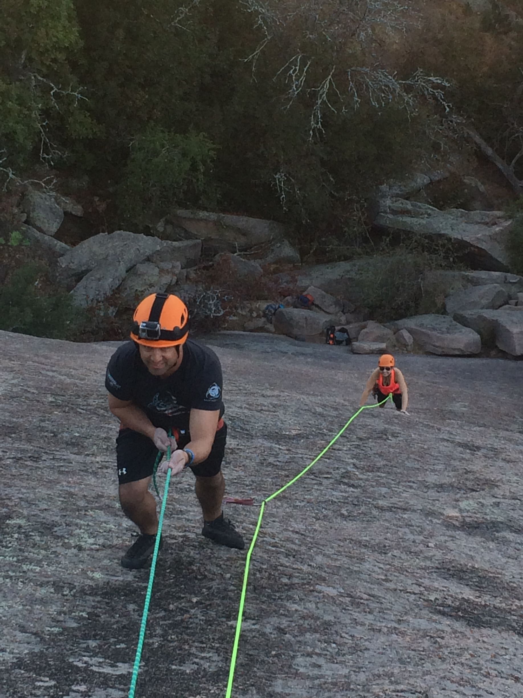 A man in a helmet is holding a rope while another man climbs a rock.