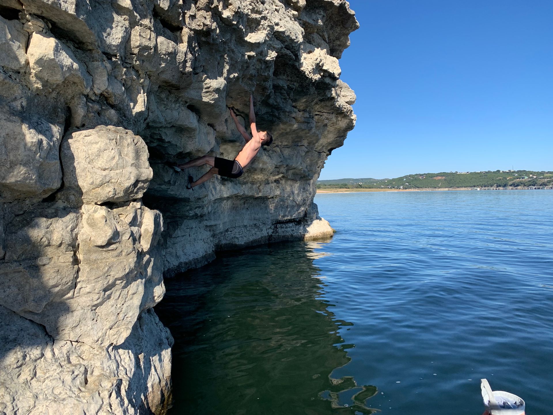 Person climbing a rocky cliff above calm blue water, with a white boat in the distance