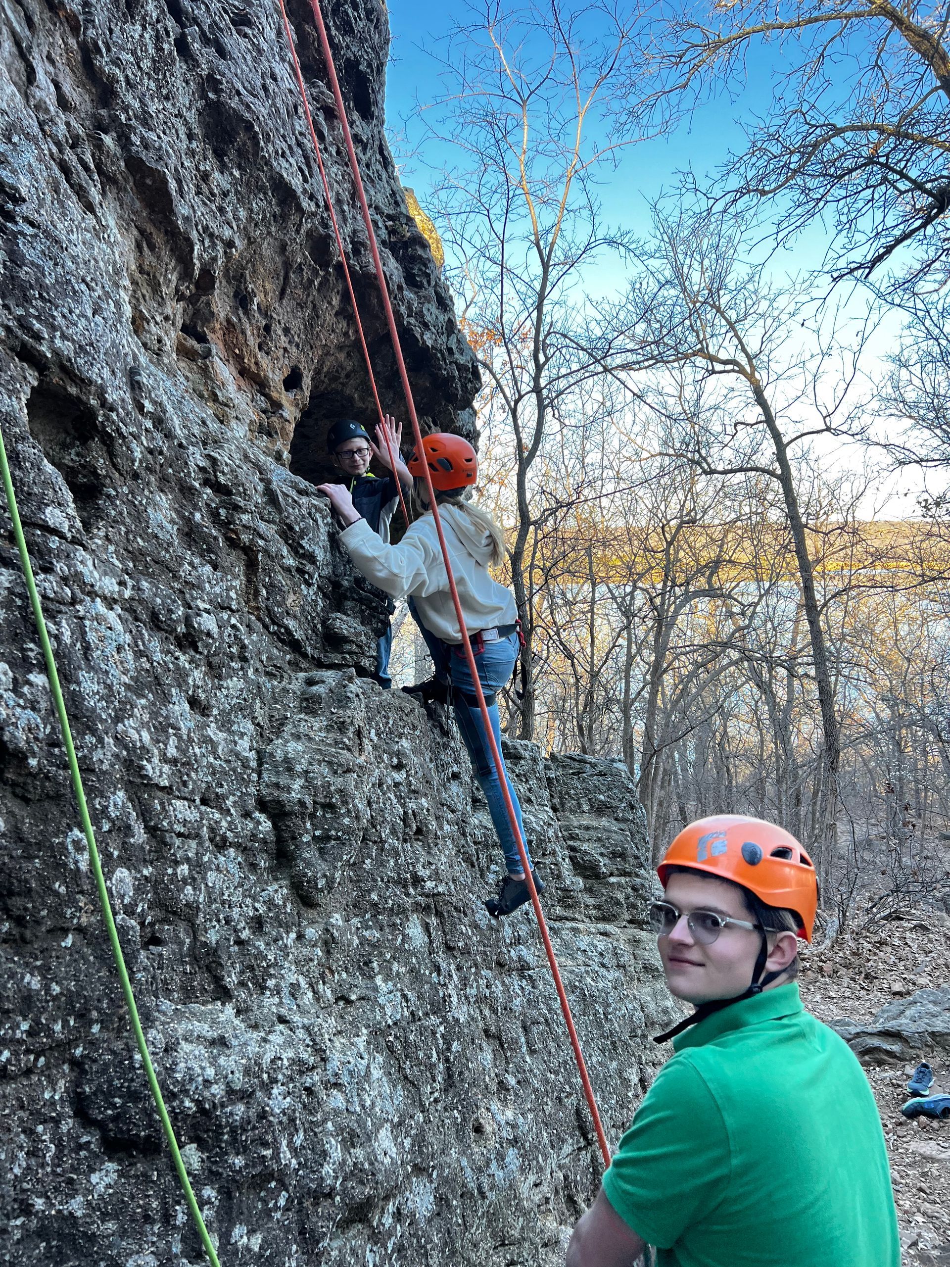 A man in a green shirt is standing next to a man climbing a rock wall.