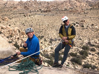 Two climbers wearing a helmets and gear on a rocky peak, one gives thumbs up
