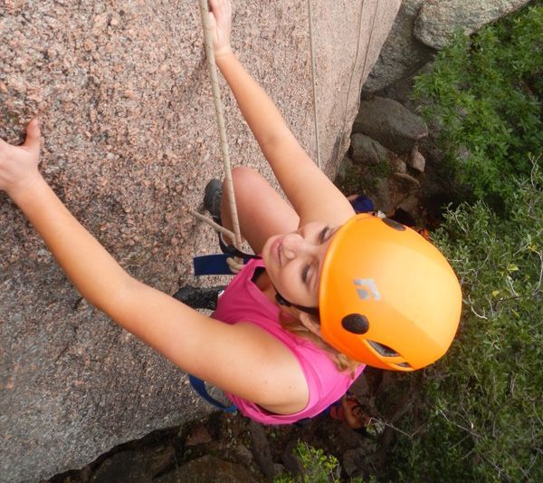 Woman rock climbing, wearing orange helmet, pink shirt, blue harness.