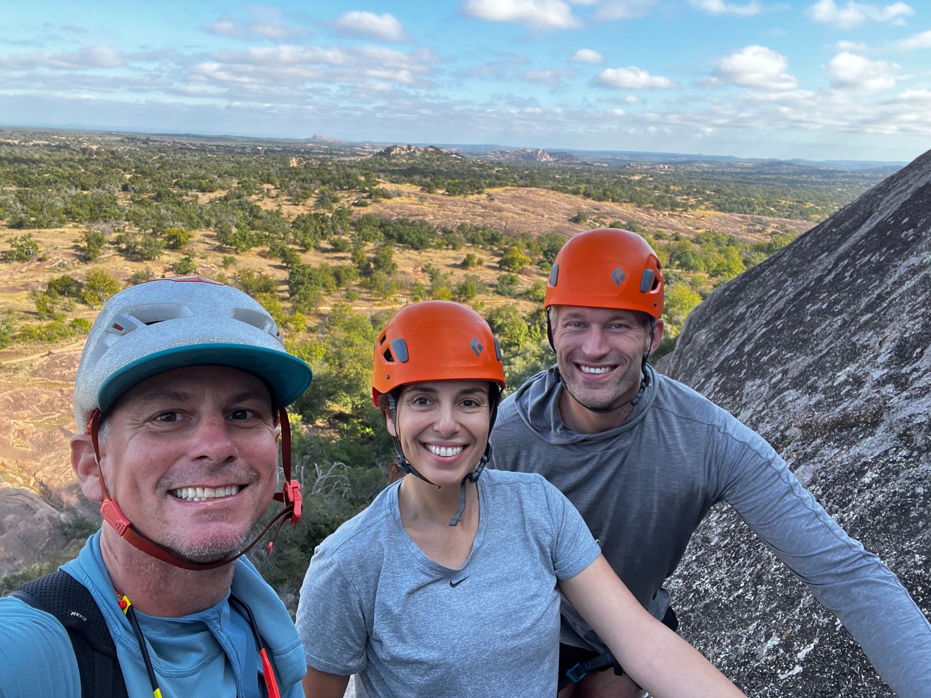 Three smiling people in helmets on a rock, overlooking a landscape.