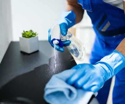 A man in blue overalls and blue gloves is cleaning a counter with a cloth and spray bottle.