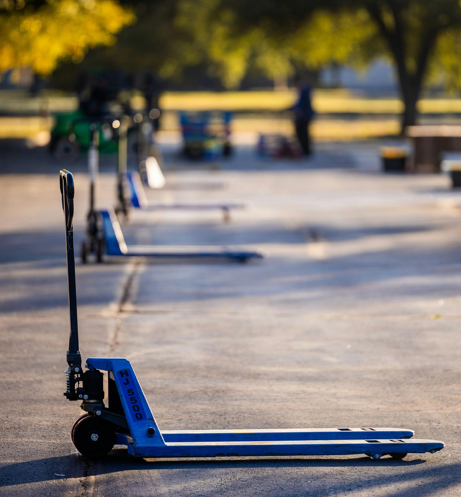 Blue pallet jacks lined up on pavement, with a blurry outdoor background.