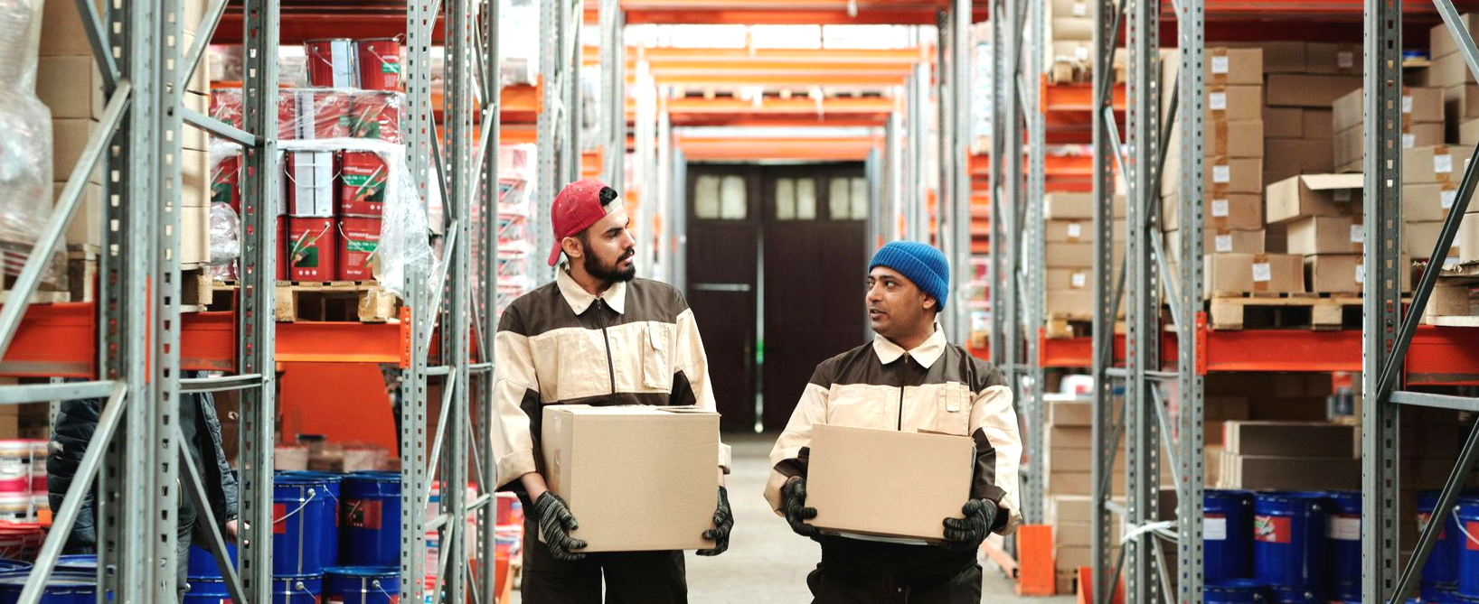 Two workers carrying boxes in a warehouse, walking down an aisle lined with shelves and goods.