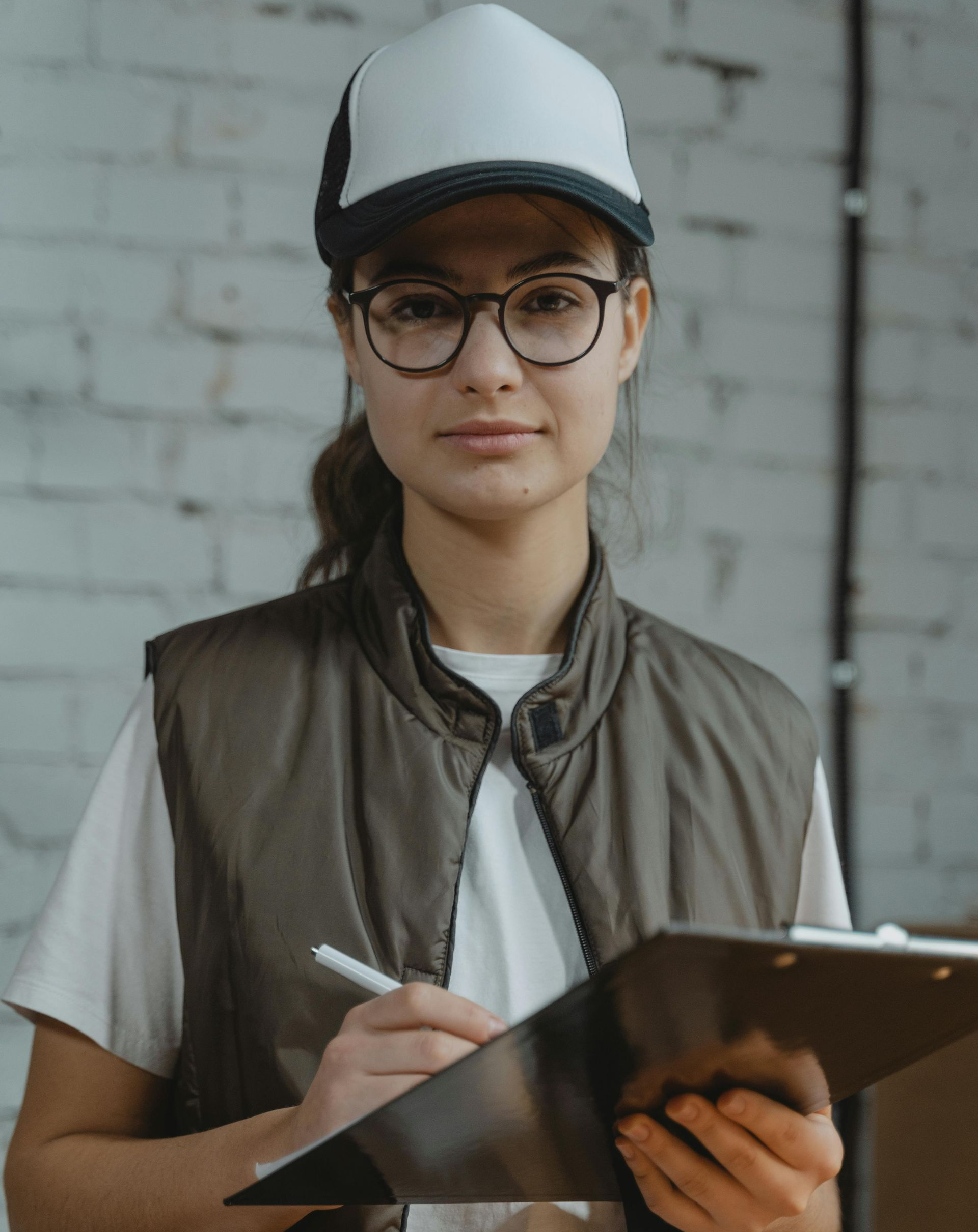 Person in cap and vest writing on clipboard, standing against a brick wall.