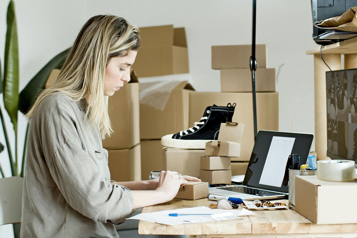 Woman packing boxes at a desk, surrounded by packages and a laptop. A sneaker sits on top.