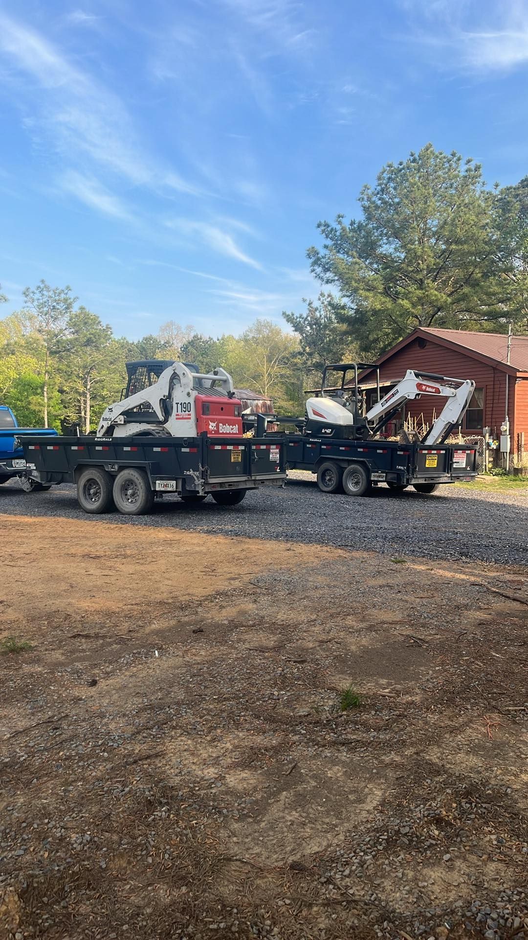 Two flatbed trucks with a white skid steer and red equipment parked on gravel near a red barn and trees.