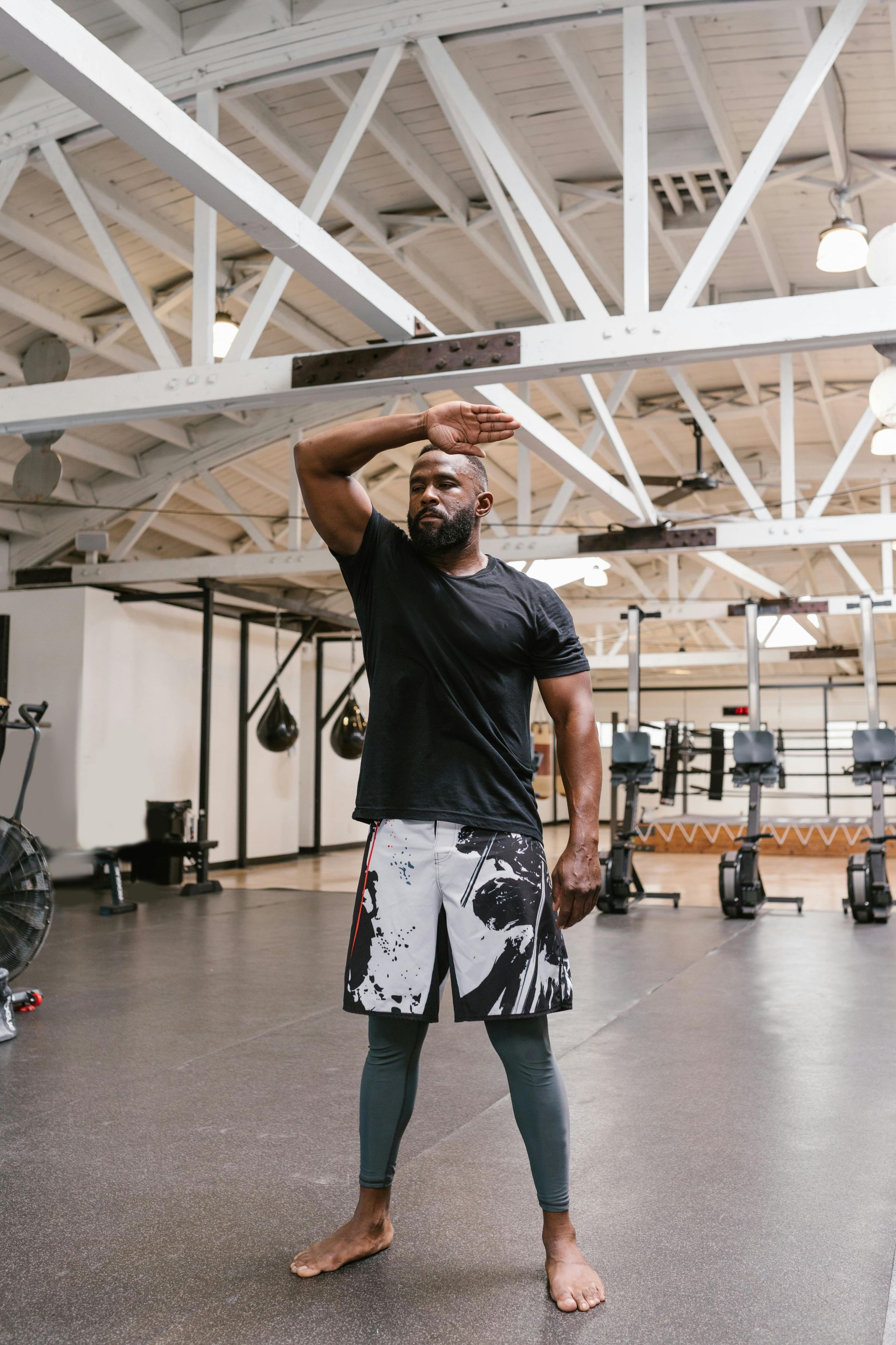 Man stretching arm in a gym, wearing black shirt, patterned shorts, and leggings. Barefoot.