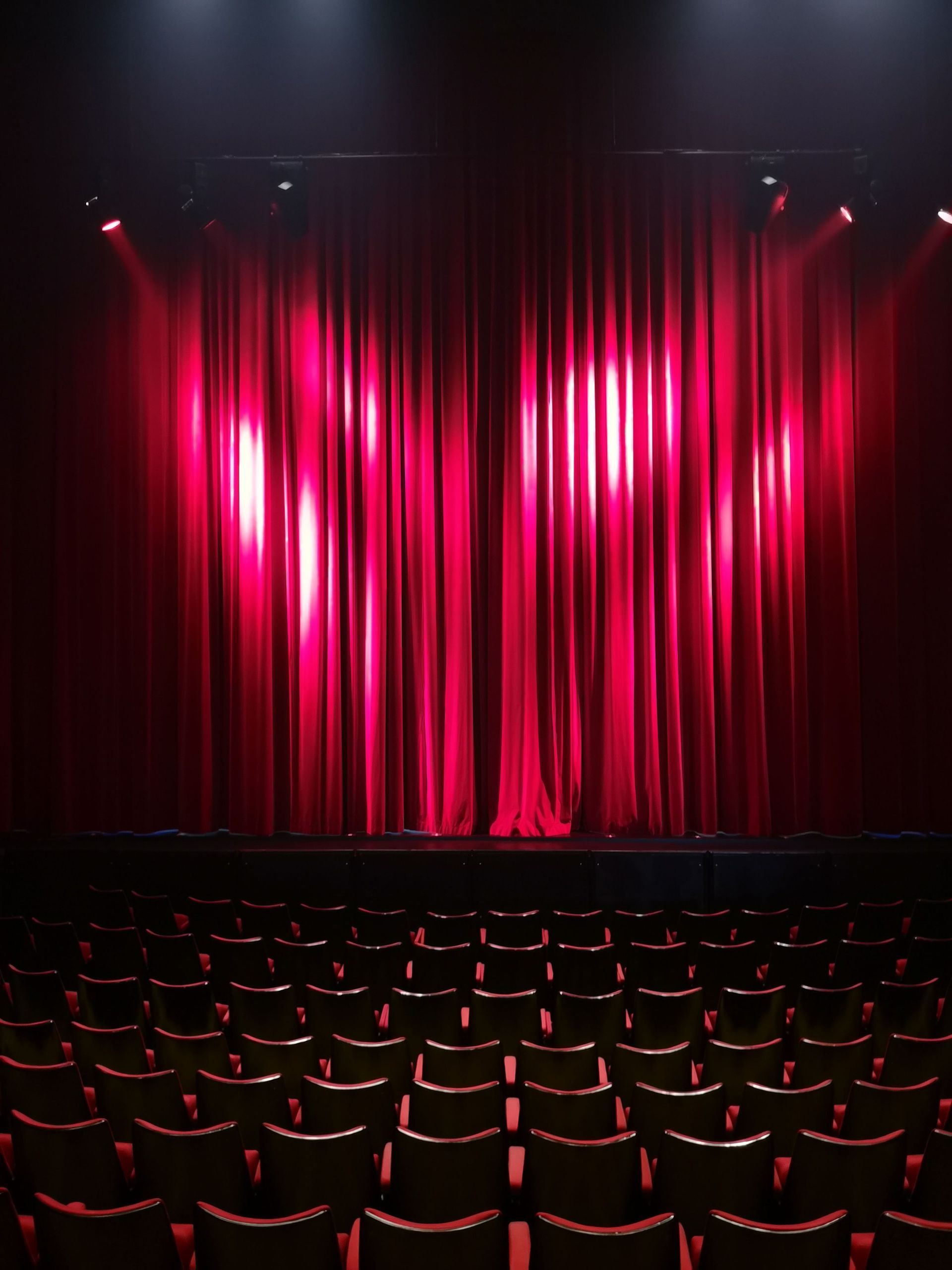 Empty theater with red velvet stage curtains lit by spotlights above rows of seats