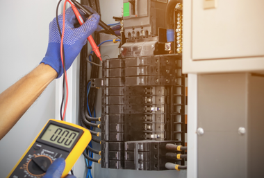 Gloved technician testing electrical wiring inside an open breaker panel with a multimeter