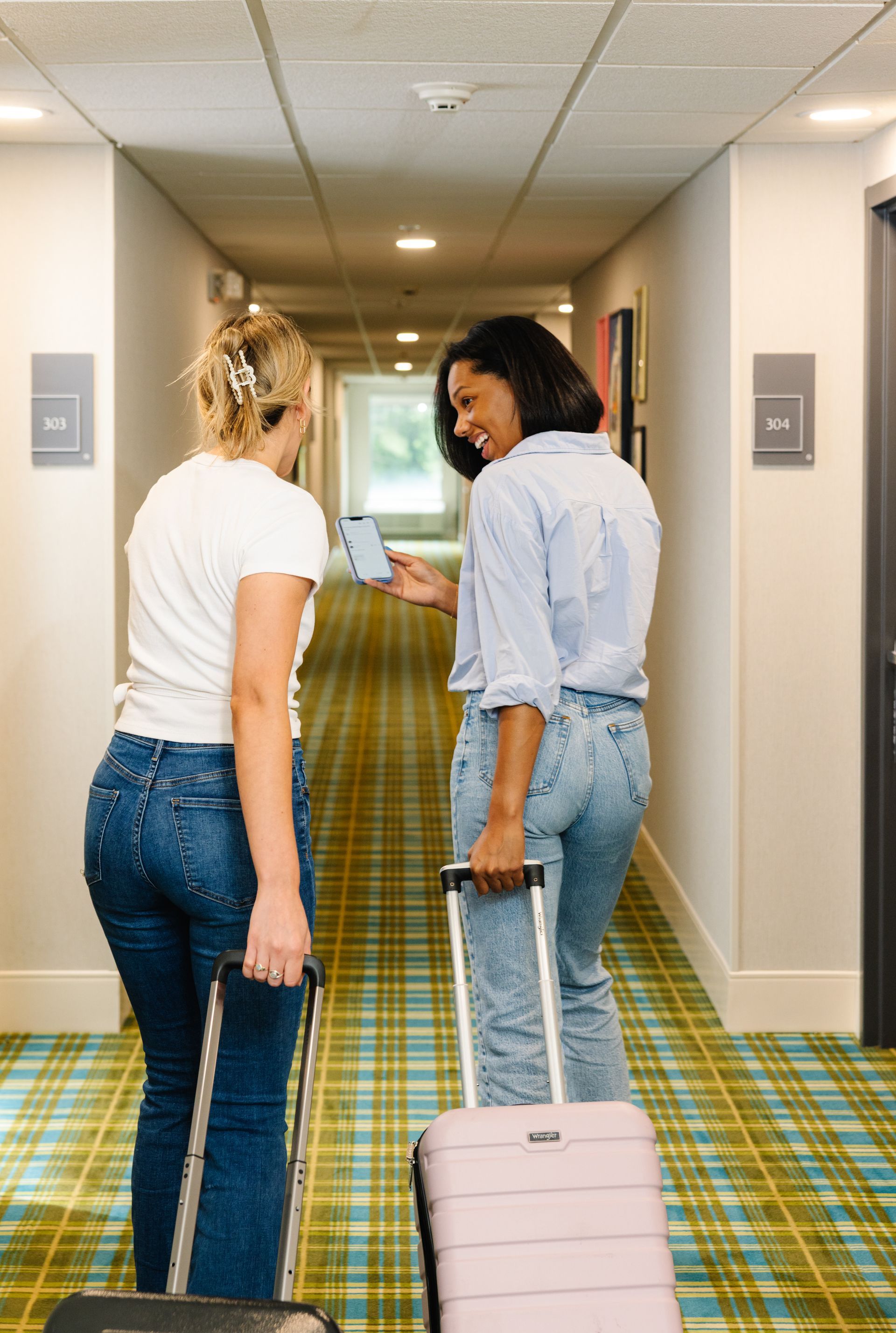 Two women are walking down a hotel hallway with luggage.