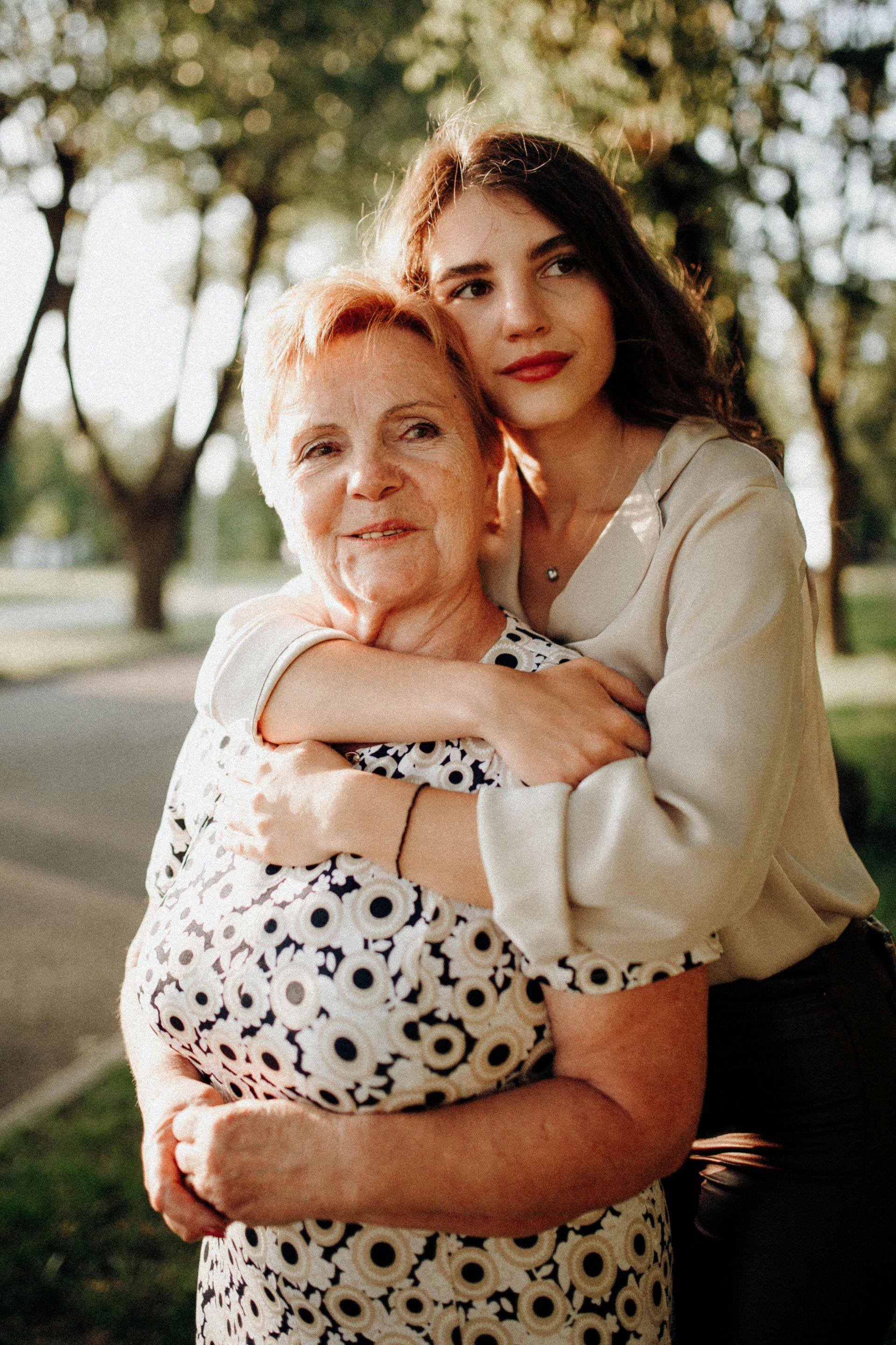Woman hugs older person outdoors; they smile, warm sunlight.