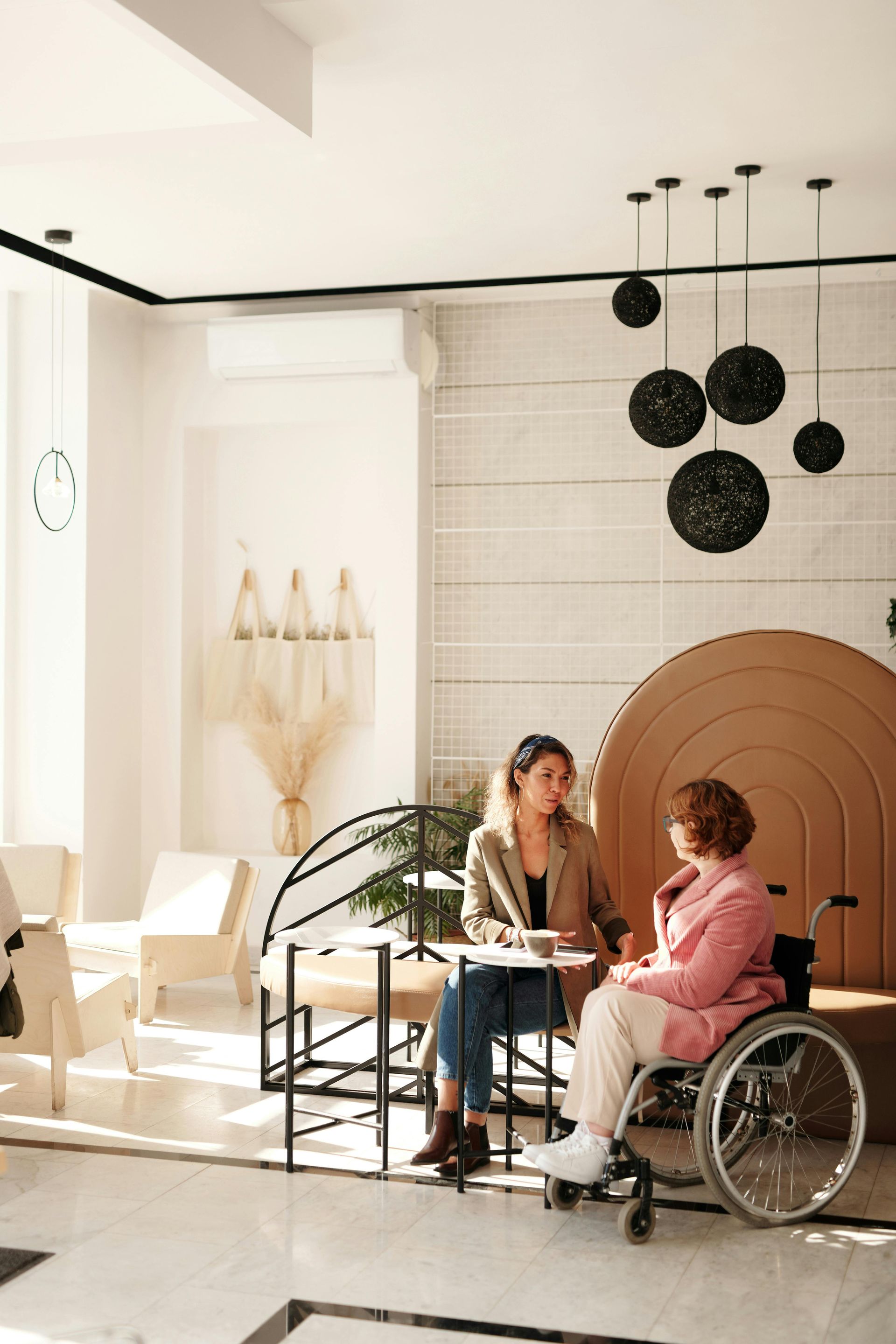Woman in wheelchair speaking with another woman at a table in a bright room.