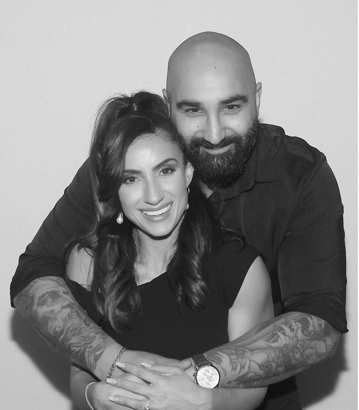 A smiling person stands in front of and embraces a bearded person with visible arm tattoos; monochrome portrait.