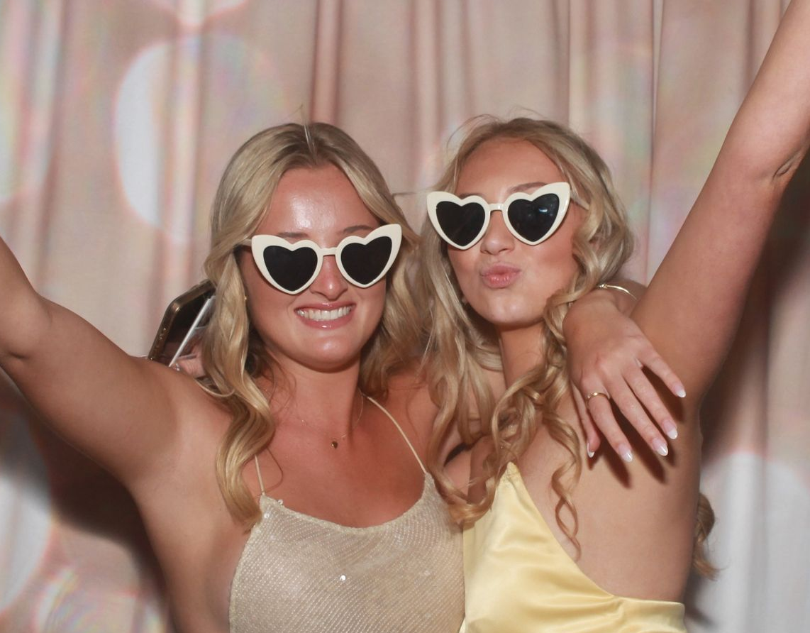 Two women wearing heart-shaped sunglasses, arms raised, smiling. Background: blurred light pink curtains. School Formal Photo Booth