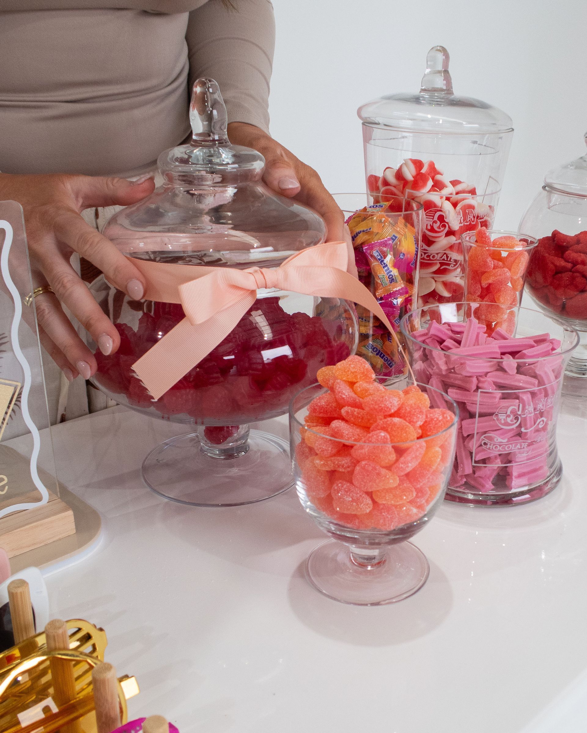 Person arranging a candy buffet with various glass jars filled with red, pink, and orange candies.