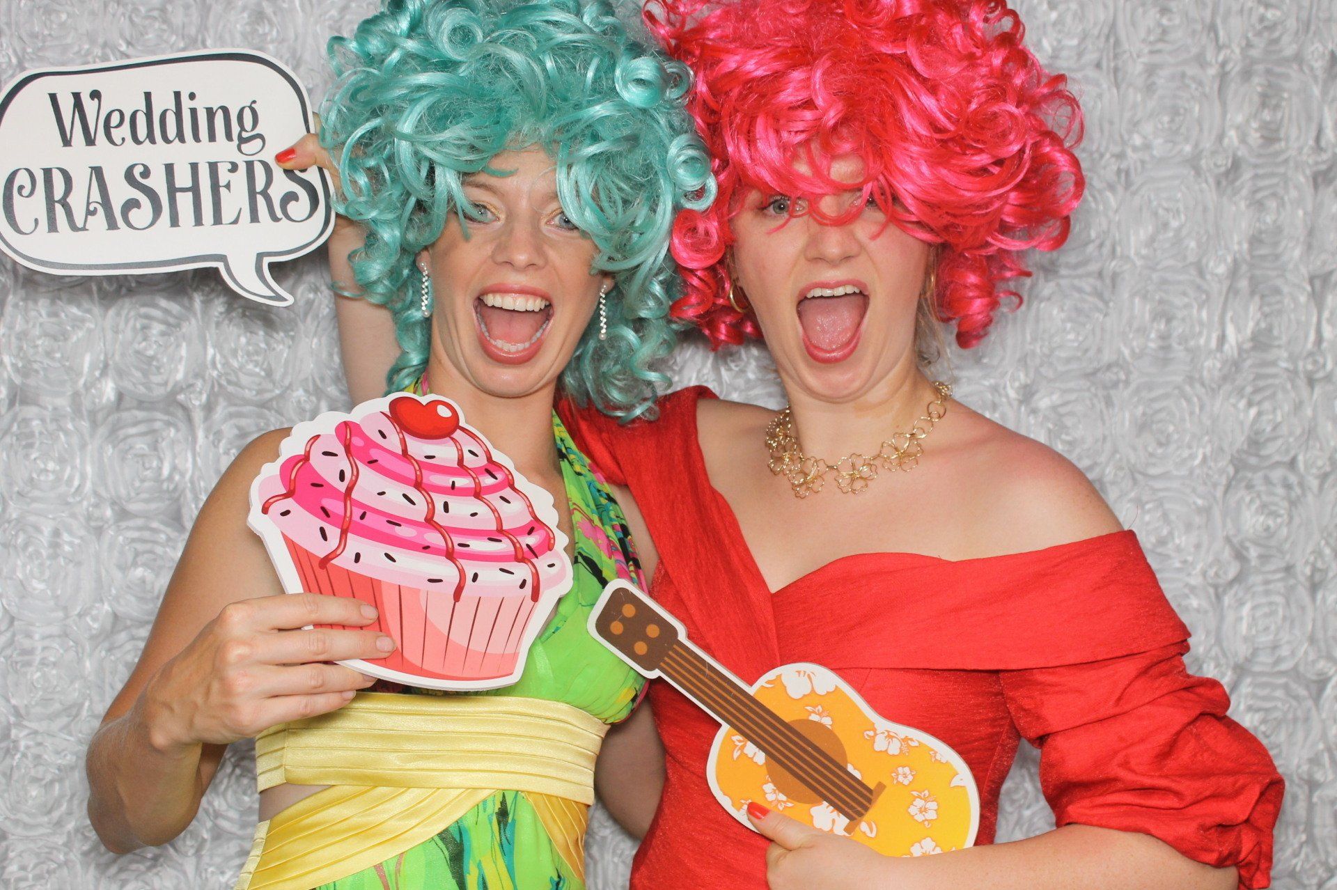 Two women with wigs, holding props, posing in a photo booth.